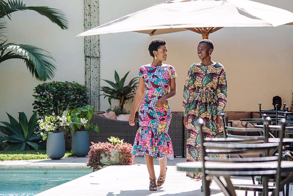 Two women wearing a fitted floral midi dress and a layered peplum dress from Queen of Chitenge Fashion’s 2023 collection